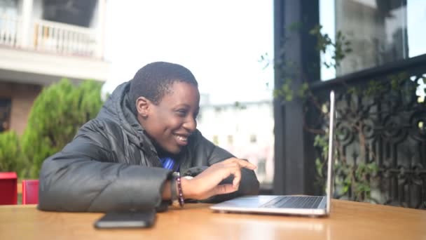 Glücklich lächelnde afrikanische Studentin mit schwarzem Kurzhaarschnitt in schwarzer Daunenjacke und blauen Kopfhörern unterhält sich auf dem Laptop im Straßencafé. Vielfalt. Fernarbeit, Fernstudium. — Stockvideo
