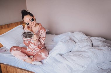 Kozmetik, cilt bakımı, yüz bakımı, spa, doğal güzellik konsepti. Pembe ipek pijamalı, gülümseyen mutlu genç kadın aynayla yatakta oturuyor ve gözlerine siyah nemlendirici bantlar sürüyor. Güzellik rutini.