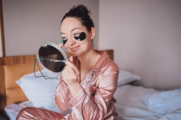 Cosmetology, skin care, face treatment, spa, natural beauty concept. Smiling happy young woman in pink silk pajamas sits on bed with mirror applying black moisturizing patches on eyes. Beauty routine. — 图库照片