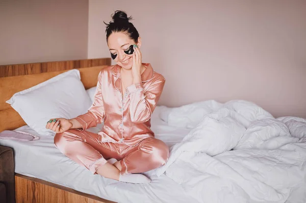 Cosmetology, skin care, face treatment, spa, natural beauty concept. Smiling happy young woman in pink silk pajamas sits on bed with black moisturizing patches on her eyes. Beauty routine. — Fotografia de Stock