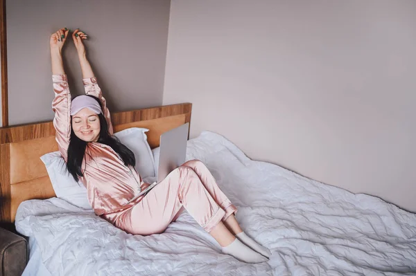 ピンクのシルクのパジャマとアイスリーピングマスクを着た魅力的な笑顔の若い女性がベッドで一人で目を覚ます幸せなコンセプト、快適なベッドで健康な睡眠の後目を覚ますとマットレスは良い朝をお楽しみください. — ストック写真