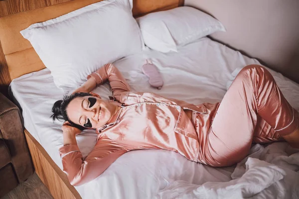 Cosmetology, skin care, face treatment, spa, natural beauty concept. Smiling young woman in pink silk pajamas lies in bed with black moisturizing patches on her eyes. Beauty routine. — 图库照片