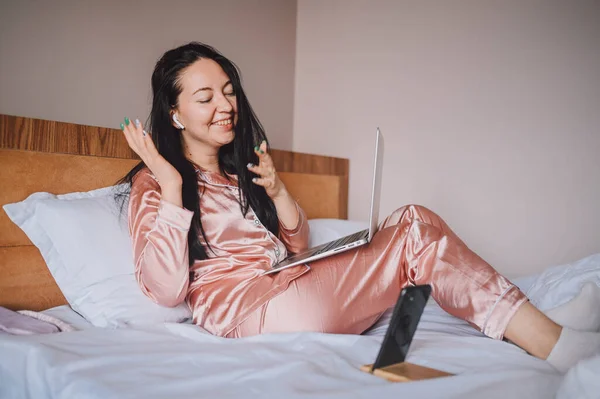 Young happy brunette woman with wireless earphones lying in bed in pink silk pajamas with smartphone and laptop computer, have online video chat. Blogger broadcasts live morning stream — ストック写真