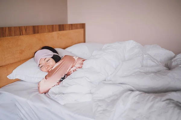 Atractiva joven sonriente en pijama de seda rosa y máscara de dormir para los ojos estirándose en la cama despertando solo concepto feliz, despierto después de dormir sano en cama cómoda y colchón disfrutar de buenos días. — Foto de Stock