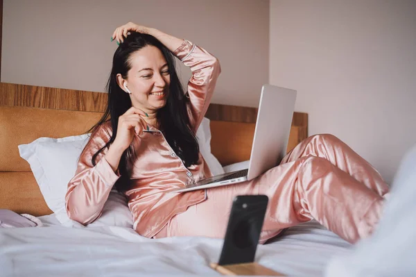 Young happy brunette woman with wireless earphones lying in bed in pink silk pajamas with smartphone and laptop computer, have online video chat. Blogger broadcasts live morning stream — Stockfoto