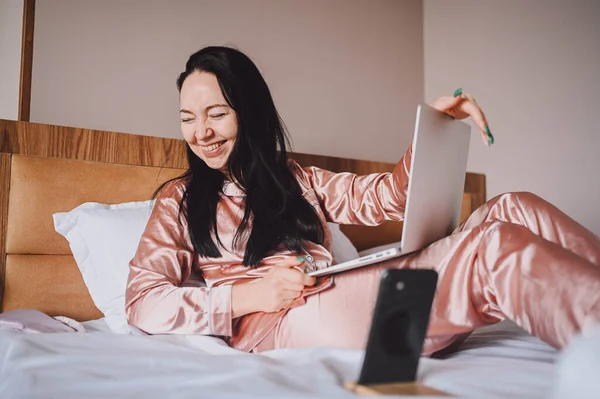Young happy brunette woman with wireless earphones lying in bed in pink silk pajamas with smartphone and laptop computer, have online video chat. Blogger broadcasts live morning stream — Stockfoto