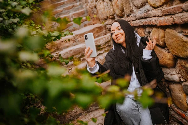 Young happy woman tourist traveling alone and making selfie with attractions by smartphone, have video chat. Blogger broadcasts live stream. Brunette lady in black down jacket and white hoodie — Stok Foto