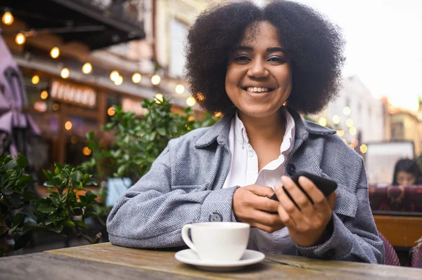 Street style portrait of attractive young happy African American woman with afro hair have video chat with smartphone outdoors in sidewalk cafe. Excited smiling female blogger broadcasts live stream. — Stok Foto