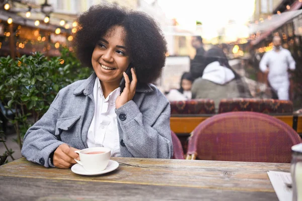 Fashion street style portrait of attractive young natural beauty African American woman with afro hair in tweed jacket speaks phone outdoors in sidewalk cafe. Happy lady with coffee cup in big city. — Stok Foto