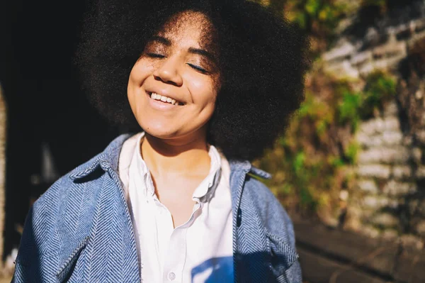 Close up Fashion street style portrait of attractive young natural beauty African American woman with afro hair in blue coat posing walking outdoors in sunny day. Happy lady with perfect teeth smile. — Fotografia de Stock