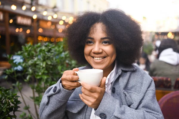 Fashion street style portrait of attractive young natural beauty African American woman with afro hair in tweed jacket posing outdoors in sidewalk cafe. Happy lady with coffee cup in big city. — Stok Foto