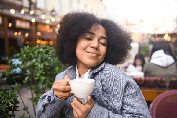 Fashion street style portrait of attractive young natural beauty African American woman with afro hair in tweed jacket posing outdoors in sidewalk cafe. Happy lady with coffee cup in big city. — Zdjęcie stockowe