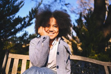 Close up Fashion street style portrait of attractive young natural beauty African American woman with afro hair in blue coat posing walking outdoors in sunny day. Happy lady with perfect teeth smile.