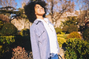 Close up Fashion street style portrait of attractive young natural beauty African American woman with afro hair in blue coat posing walking outdoors in sunny day. Happy lady with perfect teeth smile.