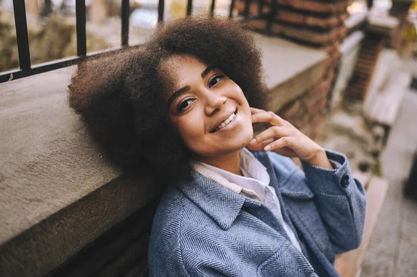 Gaya berbusana potret jalanan dari kecantikan alam wanita muda Afrika Amerika yang menarik dengan rambut afro dalam mantel biru yang berpose di luar ruangan. Selamat turis tertawa berjalan melalui pemandangan kuno bermain-main. — Stok Foto