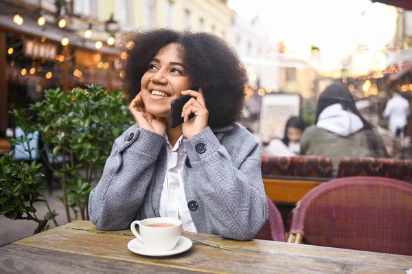 Fashion street style portrait of attractive young natural beauty African American woman with afro hair in tweed jacket speaks phone outdoors in sidewalk cafe. Happy lady with coffee cup in big city. — Stok Foto