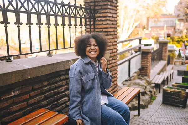 Gaya berbusana potret jalanan dari kecantikan alam wanita muda Afrika Amerika yang menarik dengan rambut afro dalam mantel biru yang berpose di luar ruangan. Selamat turis tertawa berjalan melalui pemandangan kuno bermain-main. — Stok Foto