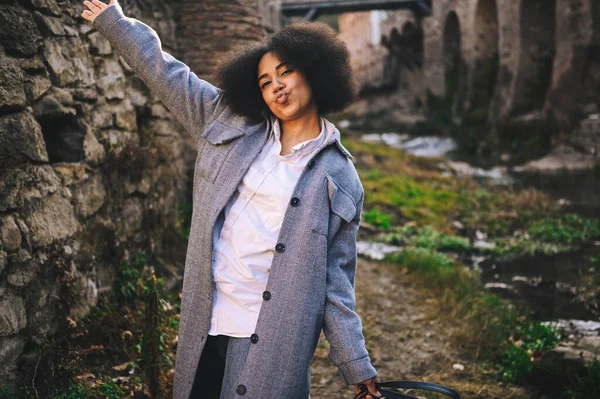 Fashion street style portrait of attractive young natural beauty African American woman with afro hair in blue coat posing outdoors. Happy tourist laughing walks through ancient sights fool around. — Stock Photo, Image