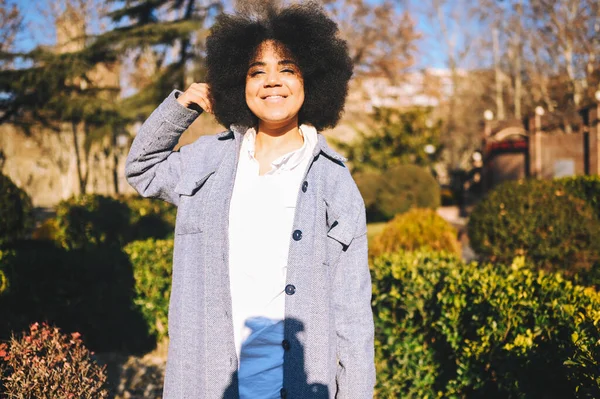 Close up Fashion street style portrait of attractive young natural beauty African American woman with afro hair in blue coat posing walking outdoors in sunny day. Happy lady with perfect teeth smile. — Stok Foto
