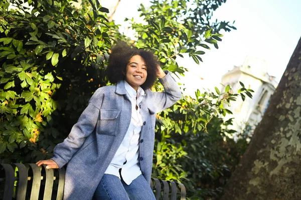 Gaya berbusana potret jalanan dari kecantikan alam wanita muda Afrika Amerika yang menarik dengan rambut afro dalam mantel biru yang berpose di luar ruangan. Selamat turis tertawa berjalan melalui pemandangan kuno bermain-main. — Stok Foto