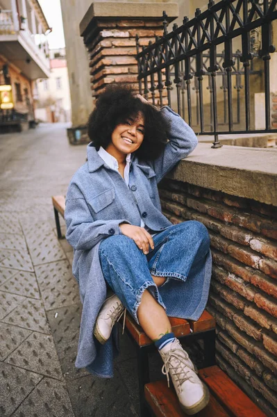 Fashion street style portrait of attractive young natural beauty African American woman with afro hair in blue coat and jeans posing outdoors. Happy tourist laughing walks around the city landscapes. — Fotografia de Stock