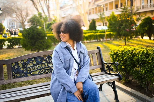 Gaya berbusana potret menarik muda kecantikan alam wanita Afrika Amerika dengan rambut afro dalam mantel biru berpose di taman alam dalam dedaunan hijau. Gigi yang sempurna tersenyum. Happy Lady dalam kacamata matahari. — Stok Foto