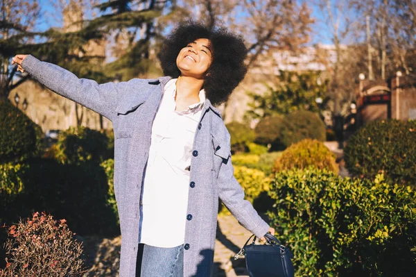 Close up Fashion street style portrait of attractive young natural beauty African American woman with afro hair in blue coat posing walking outdoors in sunny day. Happy lady with perfect teeth smile. — Stok Foto