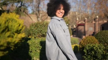 Close up Fashion street style portrait of attractive young natural beauty African American woman with afro hair in blue coat posing walking outdoors in sunny day. Happy lady with perfect teeth smile.