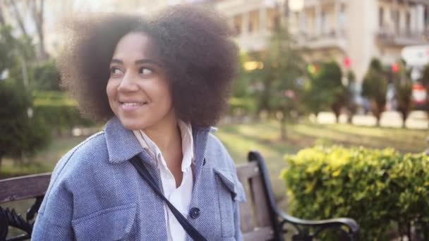 Close up Fashion street style portrait of attractive young natural beauty African American woman with afro hair in blue coat posing walking outdoors in sunny day. Happy lady with perfect teeth smile. — Stockvideo