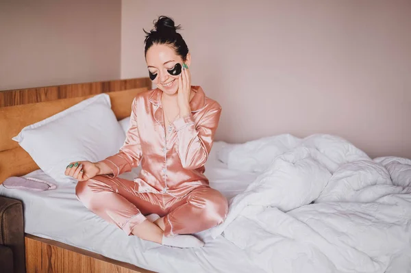 Cosmetology, skin care, face treatment, spa, natural beauty concept. Smiling happy young woman in pink silk pajamas sits on bed with black moisturizing patches on her eyes. Beauty routine. — Fotografia de Stock
