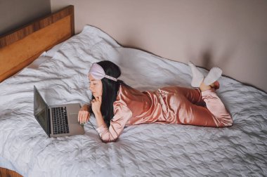 Young happy brunette woman in sleeping mask lying in bed in pink silk pajamas with laptop computer, have online video chat. Blogger or freelancer with remote work at home.