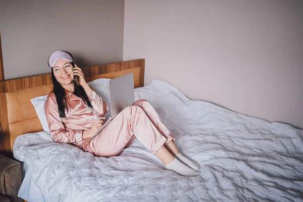 睡眠マスクの若い幸せなブルネットの女性は、ラップトップコンピュータとピンクの絹のパジャマでベッドに横たわって、電話を話し、オンラインビデオチャットをしている。自宅でリモートワークでブロガーやフリーランス. — ストック写真