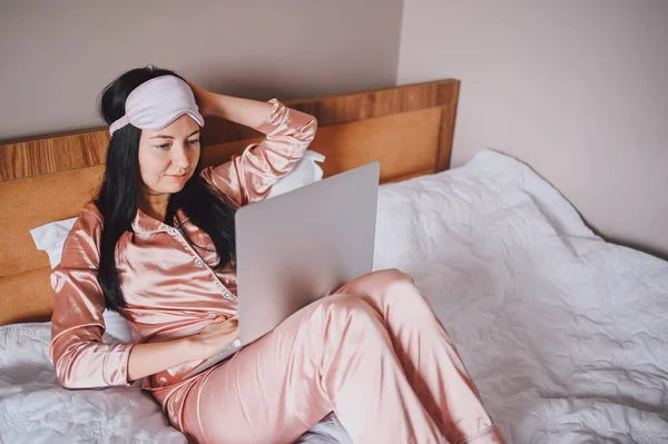 Young happy brunette woman in sleeping mask lying in bed in pink silk pajamas with laptop computer, have online video chat. Blogger or freelancer with remote work at home. — Stok Foto