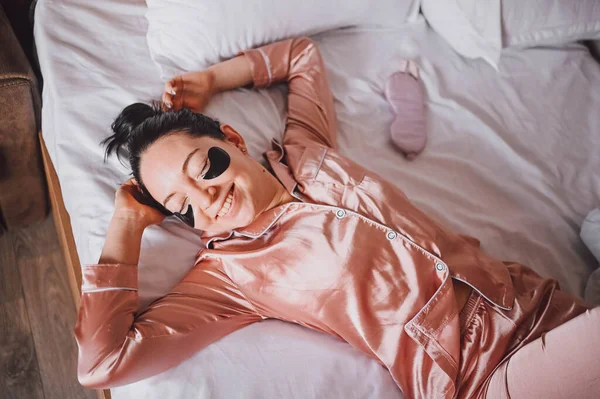 Καλλυντικά, περιποίηση δέρματος, περιποίηση προσώπου, σπα, φυσική ομορφιά. Χαμογελαστή νεαρή γυναίκα με ροζ μεταξωτές πιτζάμες ξαπλώνει στο κρεβάτι με μαύρα ενυδατικά μπαλώματα στα μάτια της. Ρουτίνα ομορφιάς. — Φωτογραφία Αρχείου