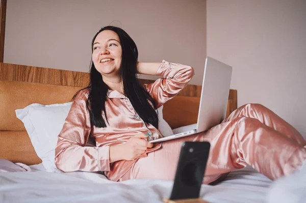 Young happy brunette woman with wireless earphones lying in bed in pink silk pajamas with smartphone and laptop computer, have online video chat. Blogger broadcasts live morning stream — ストック写真