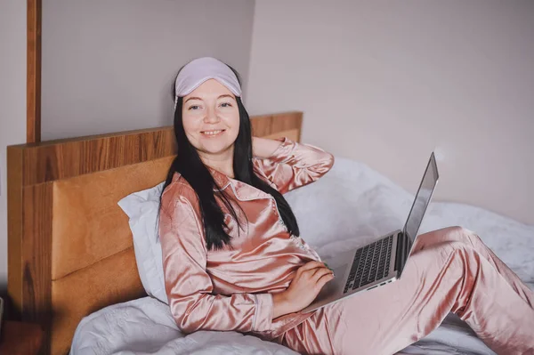 睡眠マスクの若い幸せなブルネットの女性は、ラップトップコンピュータとピンクの絹のパジャマでベッドに横たわって、オンラインビデオチャットをしている。自宅でリモートワークでブロガーやフリーランス. — ストック写真