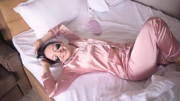 Cosmetology, skin care, face treatment, spa, natural beauty concept. Smiling young woman in pink silk pajamas lies in bed with black moisturizing patches on her eyes. Beauty routine. — Vídeo de Stock