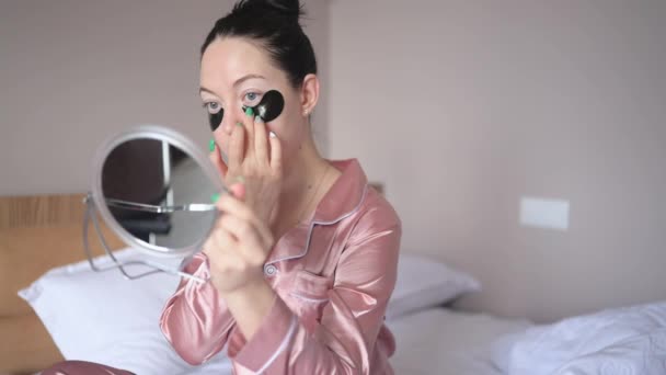 Cosmetology, skin care, face treatment, spa, natural beauty concept. Smiling happy young woman in pink silk pajamas sits on bed with mirror applying black moisturizing patches on eyes. Beauty routine. — Stockvideo