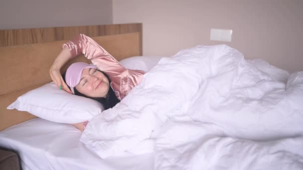 Ελκυστική χαμογελαστή νεαρή γυναίκα με ροζ μεταξωτές πιτζάμες και μάσκα ύπνου ματιών που απλώνεται στο κρεβάτι ξυπνώντας μόνη χαρούμενη έννοια, ξύπνια μετά από υγιή ύπνο σε άνετο κρεβάτι και στρώμα απολαύστε καλημέρα. — Αρχείο Βίντεο
