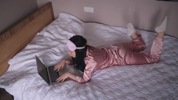 Joven morena feliz mujer en la máscara de dormir acostado en la cama en pijamas de seda rosa con ordenador portátil, tienen video chat en línea. Blogger o freelancer con trabajo remoto en casa. — Vídeo de stock