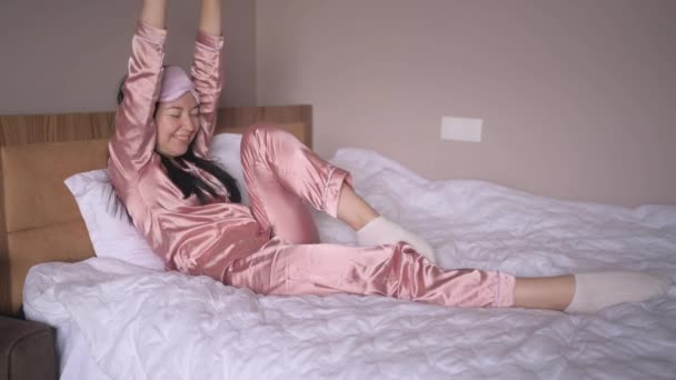 Привлекательная улыбающаяся молодая женщина в розовой шелковой пижаме и маске для сна глаз, растянувшись в постели, просыпается в одиночестве счастливой концепции, просыпается после здорового сна в удобной кровати и матрас наслаждаться добрым утром. — стоковое видео