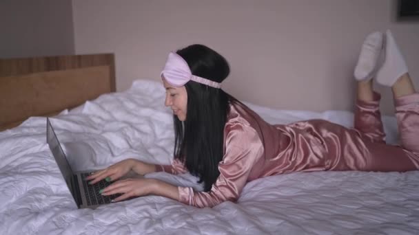 Young happy brunette woman in sleeping mask lying in bed in pink silk pajamas with laptop computer, have online video chat. Blogger or freelancer with remote work at home. — Vídeo de Stock