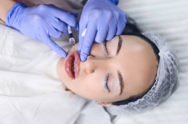 Güzellik salonunda kozmetik uzmanı hyaluronik asitle dudak büyütme ameliyatı yapıyor. Güzellik uzmanı bir iğneyle dudaklarını deliyor. Dudak şeklini arttırmak için deri altı enjeksiyonu yapan genç bir kadın.