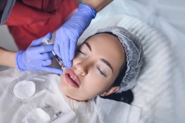 Güzellik salonunda kozmetik uzmanı hyaluronik asitle dudak büyütme ameliyatı yapıyor. Güzellik uzmanı bir iğneyle dudaklarını deliyor. Dudak şeklini arttırmak için deri altı enjeksiyonu yapan genç bir kadın.