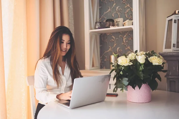 Młoda piękna azjatycka kobieta biznesu pracuje online z laptopem umieszczonym przy stole w biurze domu. Uśmiechnięta pani freelancer mają zdalną pracę siedzi w salonie z białymi różami kwiaty. — Zdjęcie stockowe