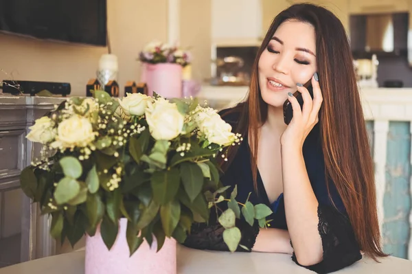 Wanita Asia yang cantik berbicara menggunakan telepon duduk di meja di ruang tamu interior yang cerah mengagumi bunga mawar putih buket. Senyum wanita muda senang menerima hadiah kejutan. — Stok Foto
