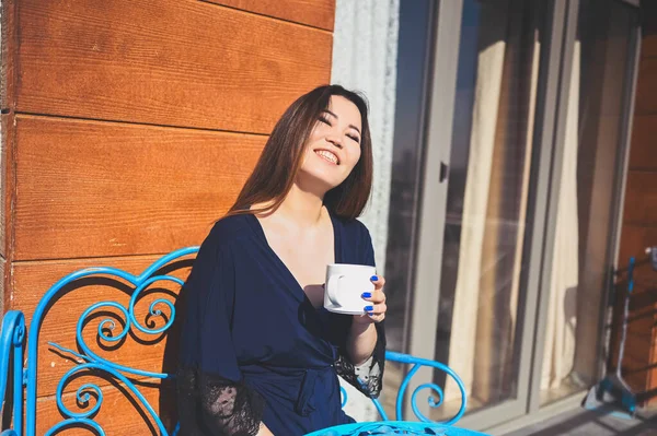 Wanita cantik Asia tersenyum menikmati kopi atau teh di balkon cerah. Wanita muda yang bahagia dengan jubah mandi biru berjemur di teras saat musim panas. — Stok Foto