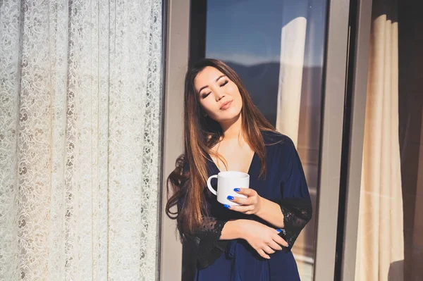 Wanita cantik Asia tersenyum menikmati kopi atau teh di balkon cerah. Wanita muda yang bahagia dengan jubah mandi biru berjemur di teras saat musim panas. — Stok Foto