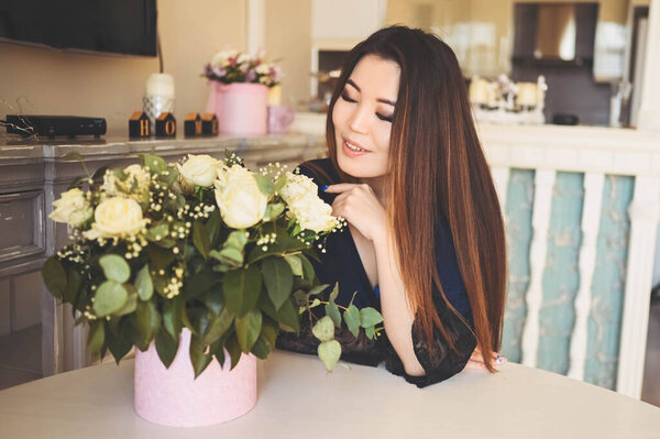 Beautiful Asian woman sits by the table in living room bright interior admires white roses flowers bouquet. Smiling young lady happy to receive a surprise gift. Royalty Free Stock Photos
