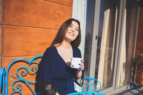 Beautiful Asian smiling woman enjoying coffee or tea on sunny balcony. Young happy lady in blue bathrobe chilling sunbathing on terrace at summertime. Royalty Free Stock Images